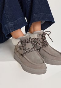 Szare zamszowe botki do kostki z puszystym białym wykończeniem, wzorzystymi sznurowadłami i grubymi, teksturowanymi podeszwami. Projekt charakteryzuje się okrągłym czubkiem i miękką cholewką.