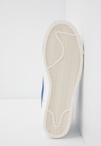 Semelle de basket blanche avec un motif en chevrons, présentant des accents bleus sur le côté et un bord lisse légèrement surélevé.