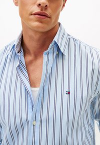 Uomo che indossa una camicia azzurra chiaro a righe verticali con piccoli ricami sul petto a sinistra, parzialmente sbottonata sopra una maglietta bianca.