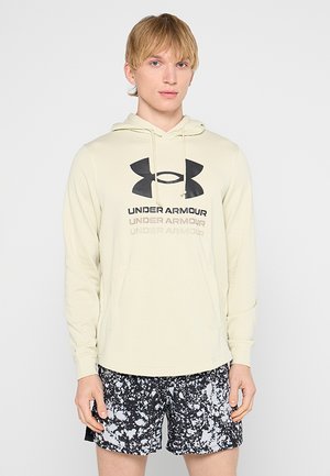 Jeune homme aux cheveux blonds courts portant un sweat à capuche beige clair Under Armour et un short à motifs noir et blanc, debout devant un fond uni.