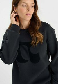 Svart sweatshirt med en mjuk textur, med en stor matta logotypdesign i mitten. Ärmarna är något oversized.