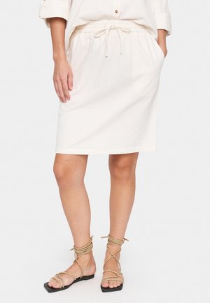 Mujer con una falda crema hasta la rodilla con bolsillos y sandalias planas beige con tiras, de pie sobre un fondo blanco.