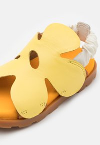 Keltainen nahkasandaali, jossa on kukkamallinen leikkaus, oranssi sisäpohja ja valkoinen joustava takahihna turvalliseen istuvuuteen.