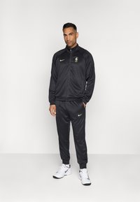 Črn trenirni komplet, ki vključuje jakno z zadrgo in logotipom na prsih ter usklajene hlače. Opojena z svetlo zelenim Nike logotipom in obuta z belimi športnimi copati.