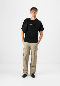 Schwarzes Baumwoll-T-Shirt mit der Aufschrift "atelier cloet." in goldener Schrift, kombiniert mit beigen Cargo-Hosen mit mehreren Taschen und schwarzen Sneakers.