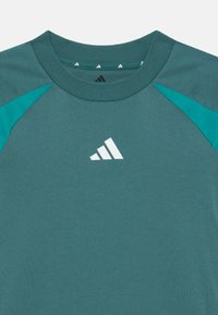 Camisa de manga corta en color verde azulado con mangas raglán. Presenta un logo de adidas en blanco en el pecho y un cuello redondo texturizado.