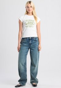 White, short-sleeve T-shirt with green text reading "LOS ANGELES Melrose WEST COAST, CA," paired with wide-leg blue denim jeans and black flats.