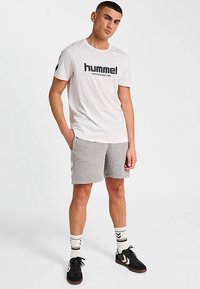 Vit T-shirt med "hummel"-logotyp, kombinerad med grå shorts. Har svarta sneakers och mönstrade strumpor. Minimalistisk design.