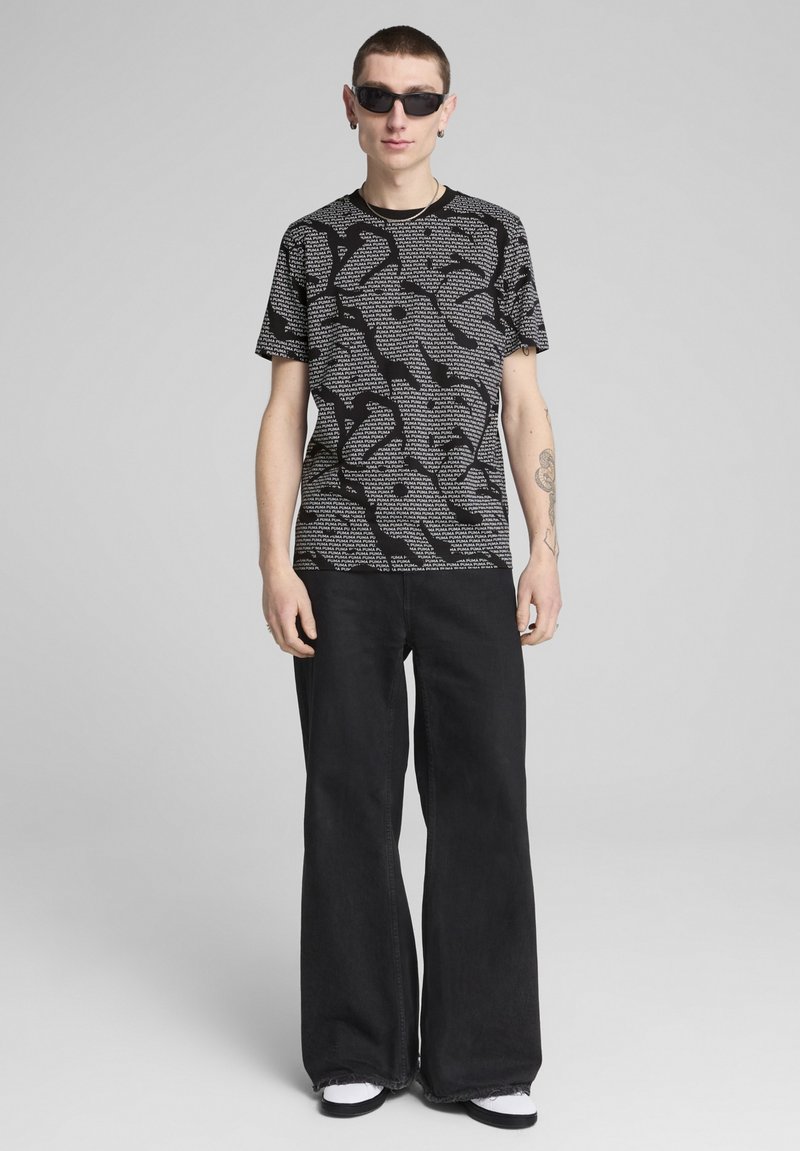 Sort grafisk t-shirt med abstrakt hvidt mønster, parret med vide sorte jeans og sorte solbriller, der viser en moderne, afslappet streetwear-stil.