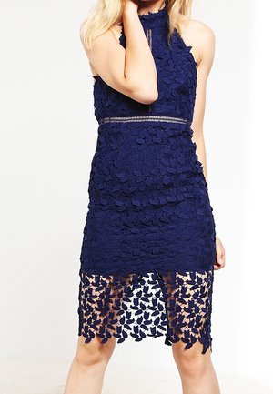 Kvinde iført en ærmeløs marineblå blondekjole med blomsterprint og en gennemsigtig nederste kant, der står mod en hvid baggrund.