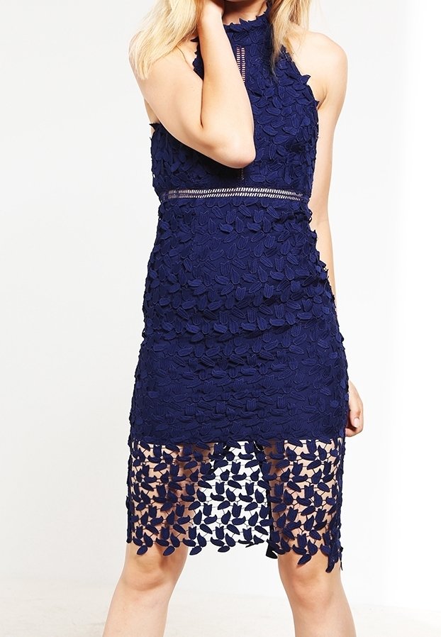 Femme portant une robe sans manches bleu marine en dentelle avec des motifs floraux et un ourlet inférieur transparent, debout devant un fond blanc.