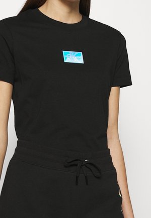 Kobieta nosząca czarną dopasowaną koszulkę z małym niebieskim prostokątnym logo Calvin Klein na środku klatki piersiowej oraz czarne spodnie ze ściągaczem.