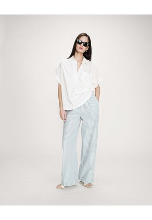 Femme portant une chemise blanche à manches courtes surdimensionnée, un pantalon large à rayures verticales bleues et blanches, des sandales blanches et des lunettes de soleil noires, debout.