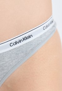 Γκρι στρινγκ με λευκή ελαστική ζώνη που φέρει την εκτύπωση "Calvin Klein" στα μαύρα. Λείος υφάσματος, στενή εφαρμογή.