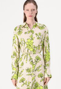 Rochie tip cămașă florală într-o nuanță crem, cu imprimeuri botanice verzi îndrăznețe. Are mâneci lungi, guler și talie strânsă cu un batic.
