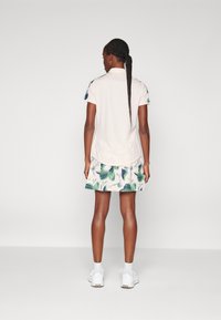 Ljusfärgad pikétröja med korta ärmar, kombinerad med en kjol med grönt lövmönster. Modellen bär vita sneakers.