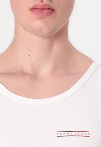 Primer plano de la parte inferior de la cara y el cuello de una persona que lleva una camisa blanca de Tommy Jeans con el logo en el pecho.