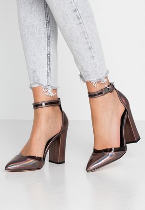 Kobieta w iryzujących szpilkach z wąskim noskiem na kostkach, w parze z jasnoniebieskimi dżinsami z postrzępionym dołem, stojąca na białej podłodze.