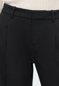 Pantalon noir tailleur avec passants de ceinture et fermeture éclair à l’avant, porté avec un top côtelé crème.