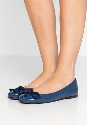 Glanzende donkerblauwe ballerina's met gedraaide touwstrikjes, gedragen op blote voeten tegen een lichte egale achtergrond.