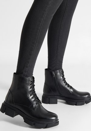 Schwarze Leder-Stiefeletten mit Schnürung vorne, seitlichem Reißverschluss, grober profilierter Sohle und glattem Finish. Kombiniert mit schwarzen Skinny-Jeans.
