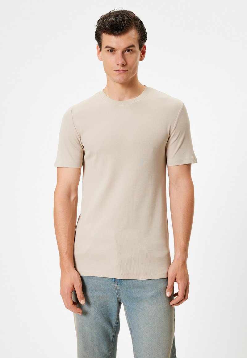 Beige ribbet t-shirt med korte ærmer, rund halsudskæring og en tætsiddende pasform, parret med lyseblå jeans. Glat tekstur, minimalistisk design.