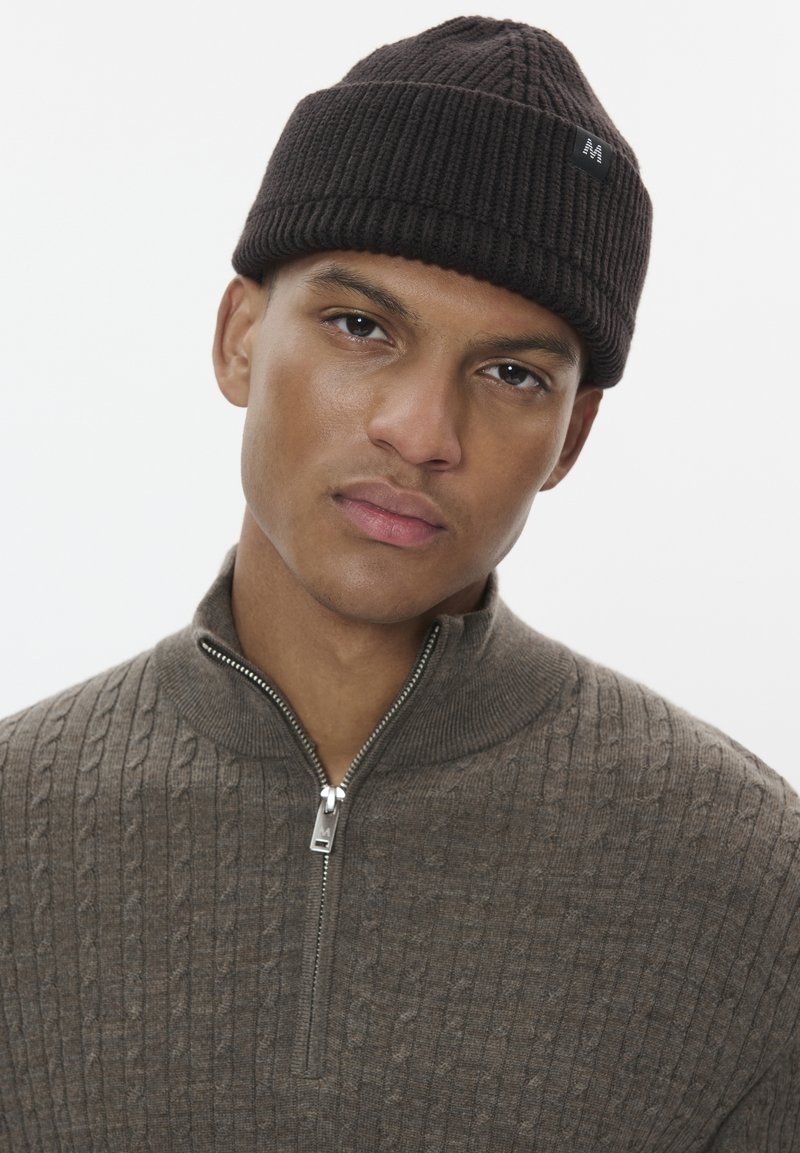 Dark brown knit beanie with ribbed texture, worn with a light brown zip-up sweater featuring a cable knit pattern and silver zipper detail.