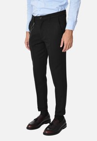 Pantaloni neri su misura con una texture liscia, dotati di un dettaglio in catena sulla tasca laterale. Abbinati a scarpe scure lucide e a una camicia azzurro chiaro.