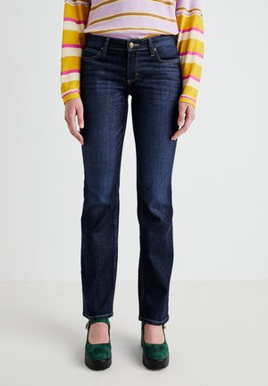Jeans de mezclilla azul oscuro con un diseño de pierna recta, que cuentan con bolsillos y detalles de costura amarilla. Combinados con zapatos de tacón cuadrado verdes.