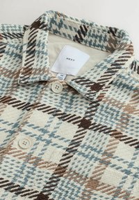 Gros plan sur une chemise à boutons en mélange de laine avec un motif à carreaux bleu, marron et crème et un col pointu.
