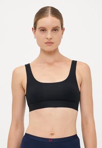 Woman with slicked-back hair wears a black sports bra and navy waistband pants, facing forward against a plain white background.