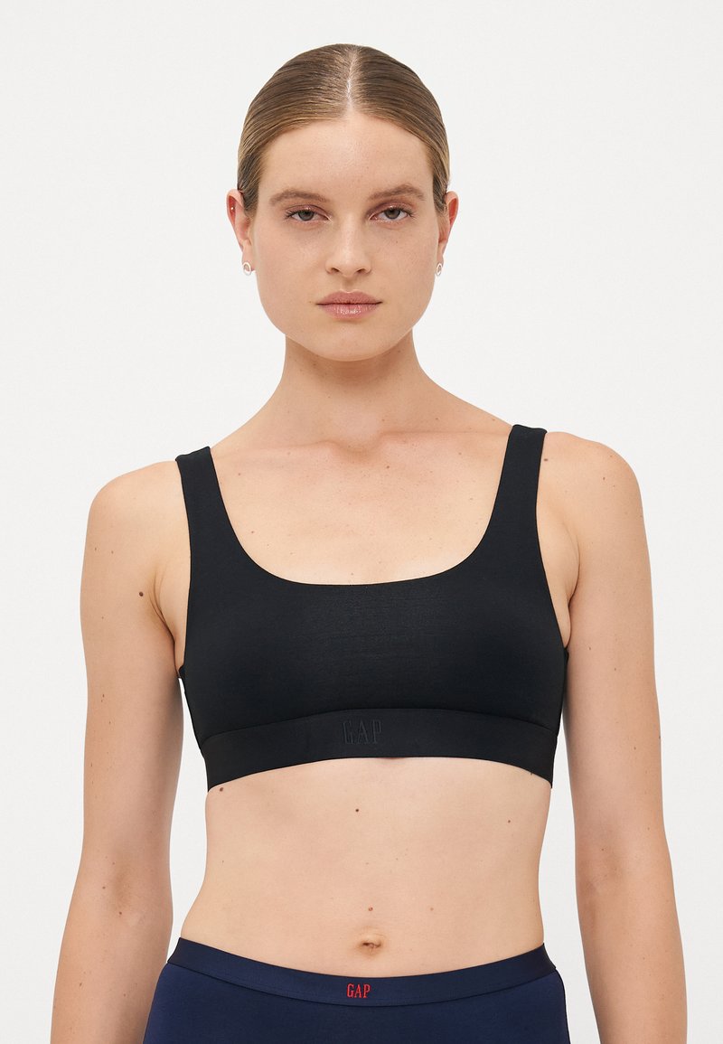 Woman with slicked-back hair wears a black sports bra and navy waistband pants, facing forward against a plain white background.