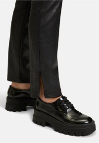Zwarte platform schoenen met een glanzende patenten afwerking, dikke rubberen zool en vetersluiting, gecombineerd met gestructureerde zwarte broeken met ritsen.