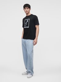 Νέος άνδρας με μαύρο graphic t-shirt, ανοιχτό μπλε φαρδιά τζιν και λευκά αθλητικά παπούτσια πόζαρε μπροστά σε ένα απλό λευκό φόντο.