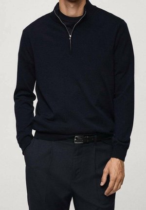 Homme portant un pull noir à fermeture éclair quart de zip par-dessus une chemise noire, avec un pantalon noir et une ceinture noire, debout avec une main dans la poche.