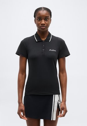 Donna in piedi che indossa una polo nera con il logo "Malbon" e una gonna nera con strisce laterali bianche e il testo "Malbon".