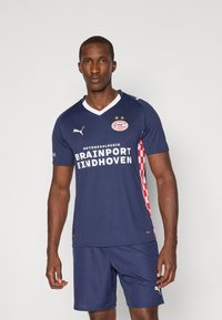 Mörkblå fotbollströja med vit V-hals, prydd med röd och vit rutig mönster på sidan, lagets logotyp och sponsortext. Matchas med blå shorts.