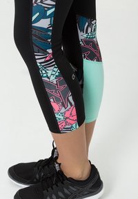 Černé sportovní legíny s barevnými květinovými vzory na stranách, s akcentem v mátově zelené barvě. Černé sportovní boty dokončují celkový vzhled.