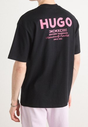 Personne portant un t-shirt noir avec un texte rose "HUGO MCMXCIII Registered Trademark Internationally Established Since 1993" dans le dos.