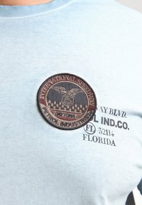 Lys blå bomulds-t-shirt med et rundt broderet mærke, der er mærket "International Speedway Daytona" samt trykt tekst nederst.