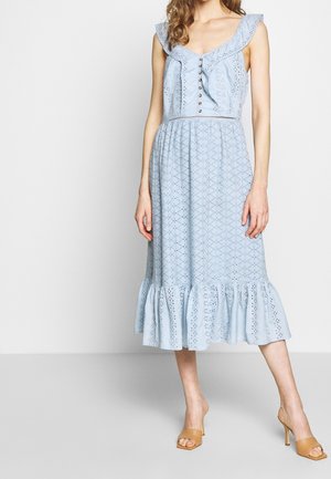 Femme portant une robe midi bleu clair sans manches en tissu à œillets avec un col à volants, des boutons sur le devant et un ourlet à volants, associée à des sandales à talons couleur camel.