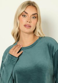 Femme blonde aux vagues lâches portant un haut en velours teal à manches longues, touchant son col près de son cou contre un fond uni.