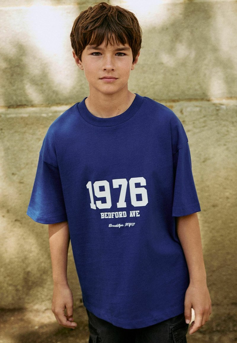 Blauwe katoenen t-shirt met witte tekst: "1976 BEDFORD AVE Brooklyn, NYC." Korte mouwen, relaxed fit, ronde hals, geen zichtbare patronen.
