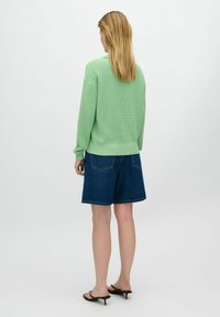 Hellgrüner, gestrickter Pullover mit langen Ärmeln und gerippten Bündchen, kombiniert mit dunklen Jeansshorts. Rückansicht; glatte Textur, lässiges Design.