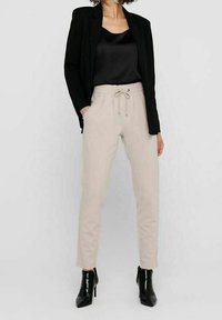 Beige tapered Hosen mit Kordelzug in der Taille, getragen mit einem schwarzen Satinoberteil und einem schwarzen Blazer. Schwarze Knöchelstiefel runden das Outfit ab.