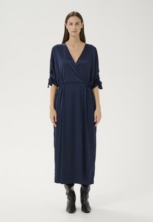 Žena stojaca v tmavomodrých saténových midi šatách s naberanými 3/4 rukávmi a čiernymi čižmami po kolená na jednoliatom bielom pozadí.