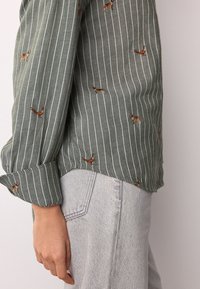 Green, striped shirt with embroidered animals, featuring long sleeves and a folded cuff. Paired with light gray, high-waisted jeans.