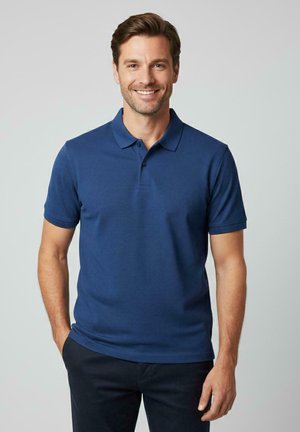 Marineblauw poloshirt met korte mouwen, drieknopenbies, gestructureerde stof, casual pasvorm, gedragen met donkere broek, model glimlacht.