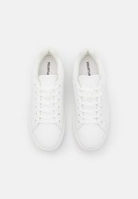 Zapatillas blancas con cordones que presentan un upper sintético suave, puntera redonda y acolchado en el cuello. Logo visible en la plantilla. Suela de goma plana.