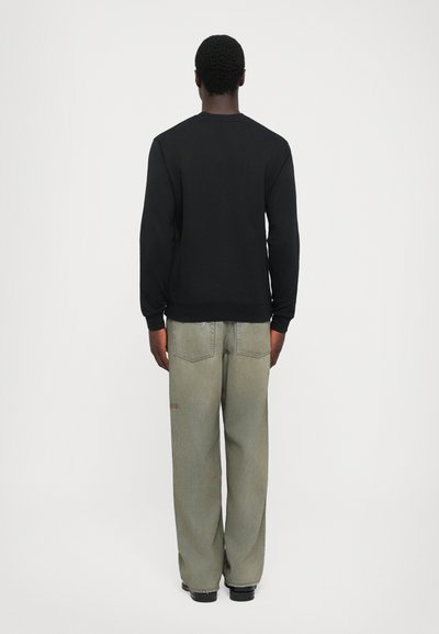 Ein Mann, der von hinten zu sehen ist, trägt ein schwarzes Langarmshirt, lockere hellgraue Jeans und schwarze Schuhe vor einem schlichten weißen Hintergrund.